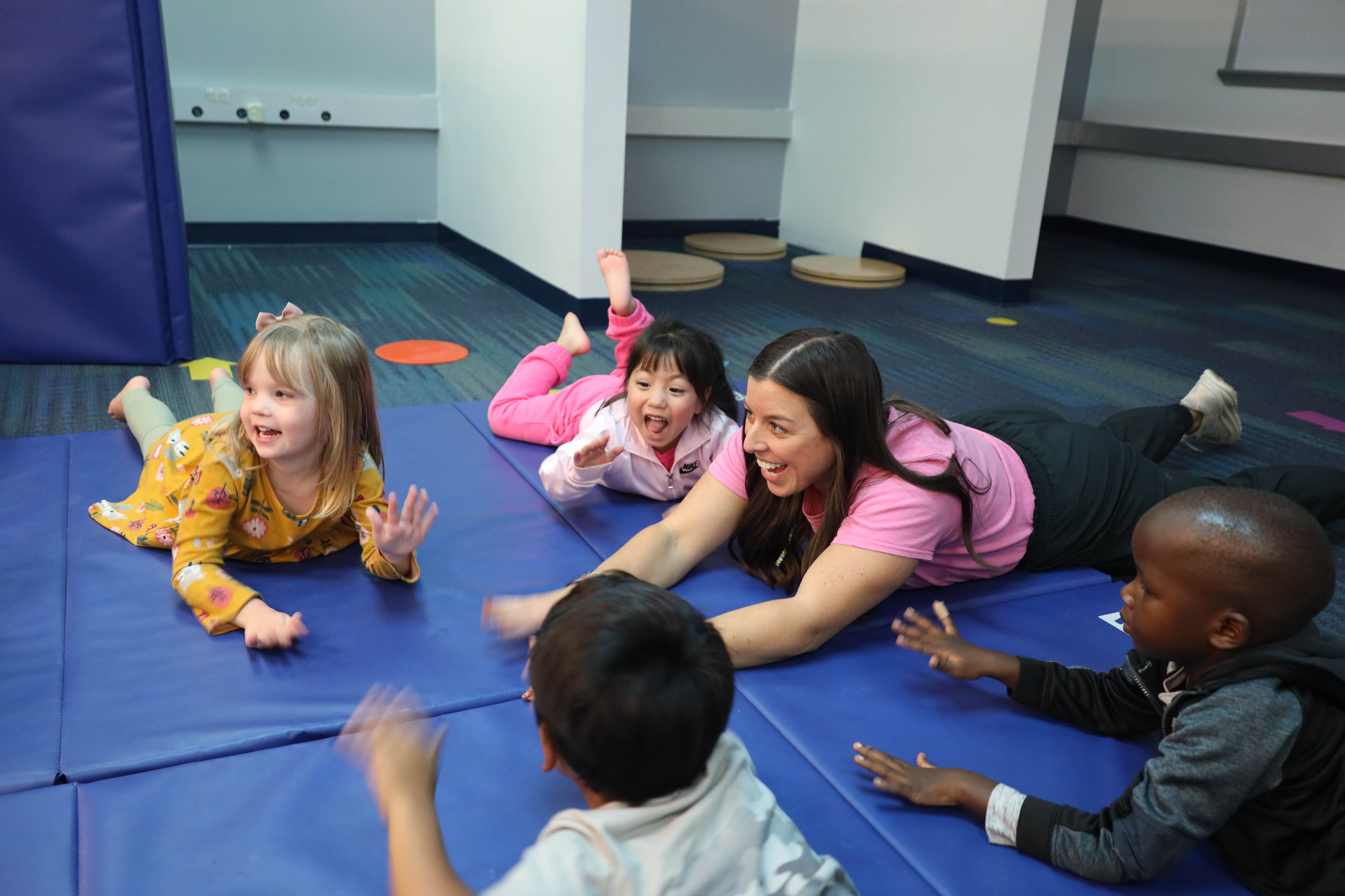 Early childhood students and a teacher kick their feet and pat their hands while laying on a mat on the floor.